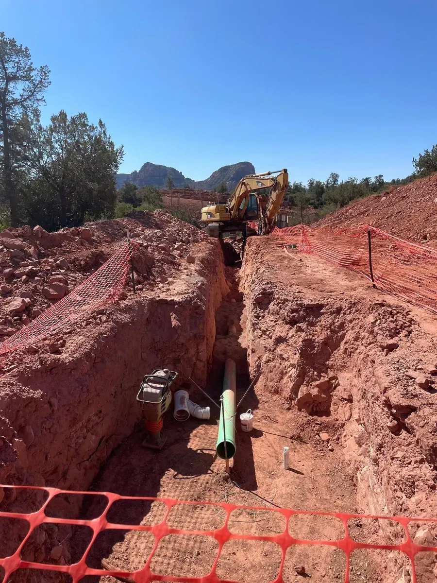 Excavator digging a deep trench in red dirt, with orange safety fencing and trees under a blue sky.