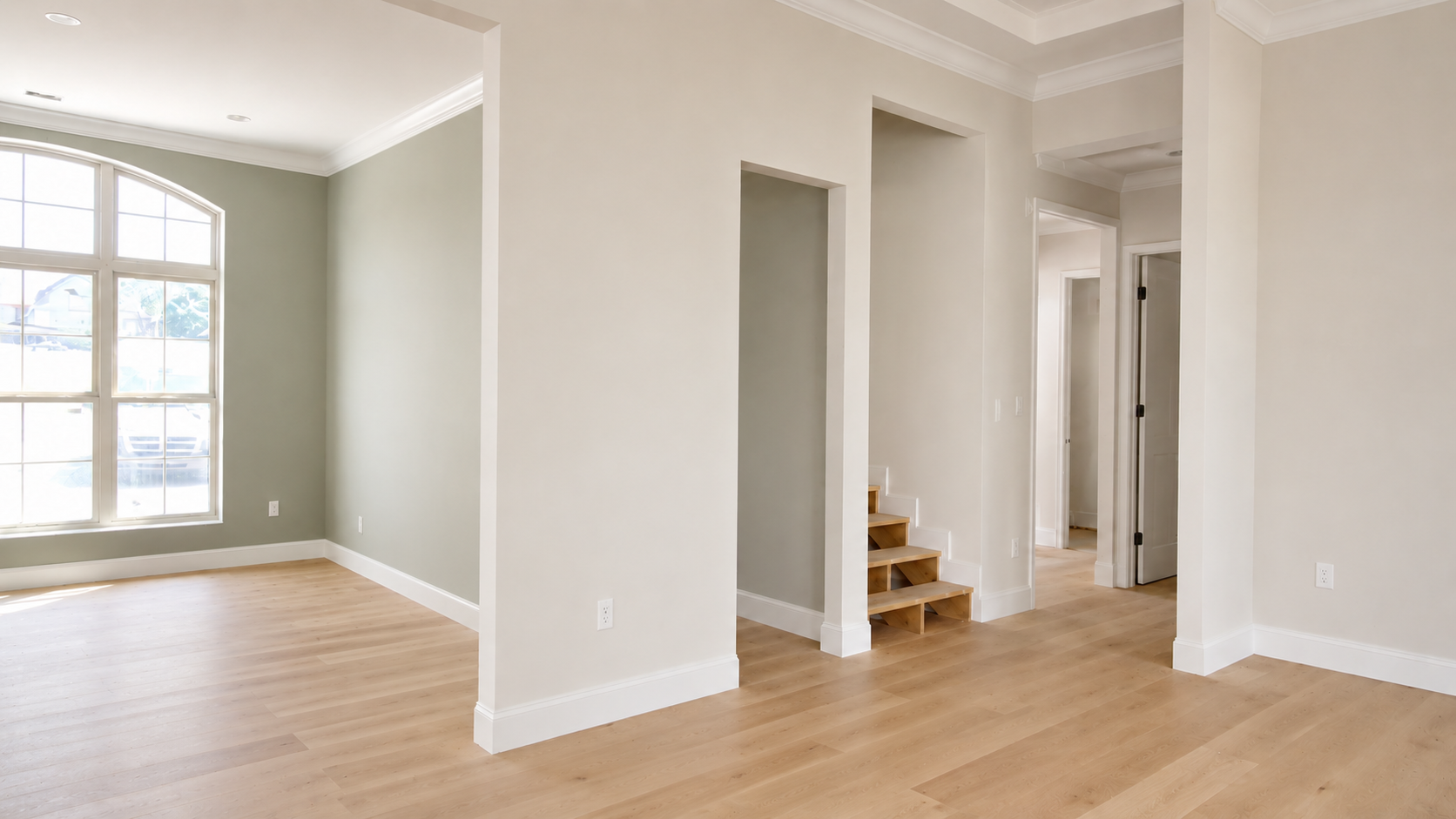 Bright empty living room with hardwood floors, large window, and a hallway with stairs.