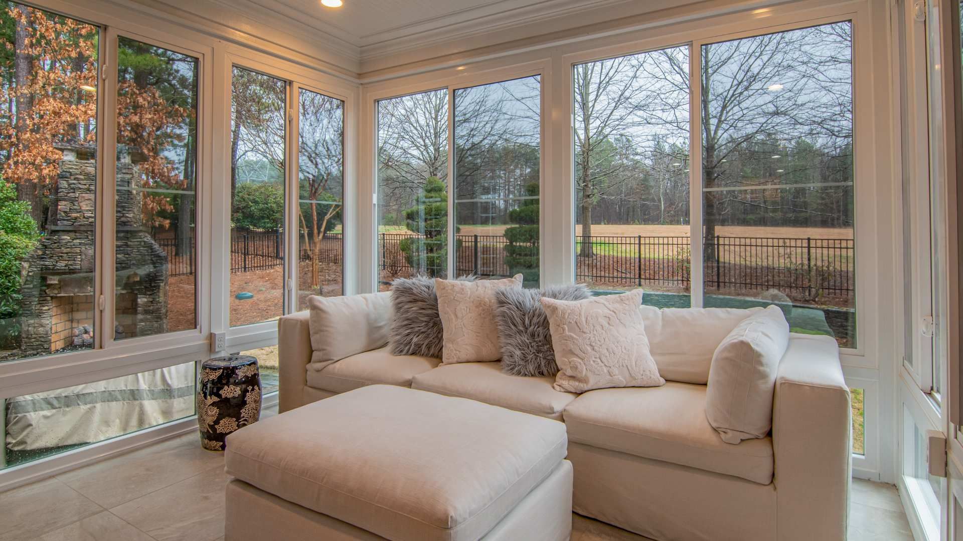 Bright sunroom with white sectional, ottoman, and large windows overlooking a wooded yard