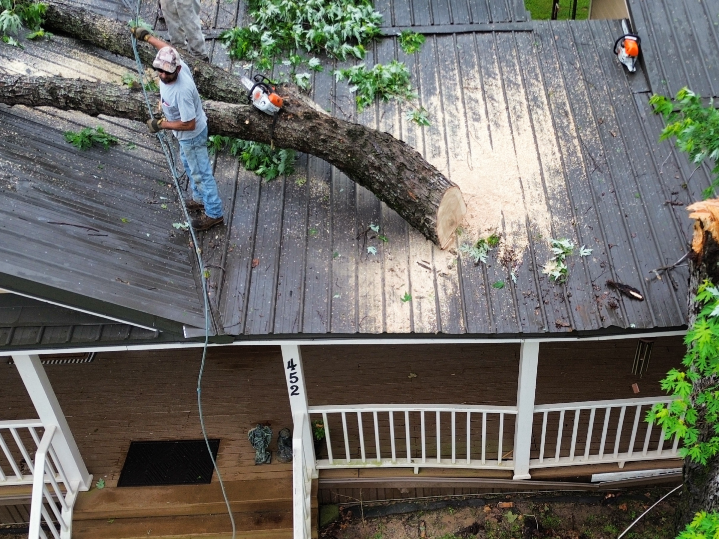 Worker trimming a large tree fallen across a house roof, with branches and a chainsaw on the shingles.