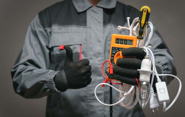 Technician holding a clamp meter with tangled wires and a test probe in a gray work uniform