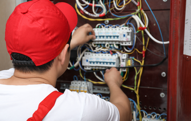 Technician wiring an electrical panel in a red cabinet, wearing a red cap and white shirt