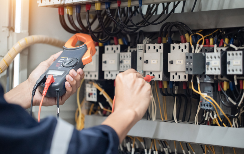 Technician testing electrical panel wiring with a clamp meter inside an open control cabinet