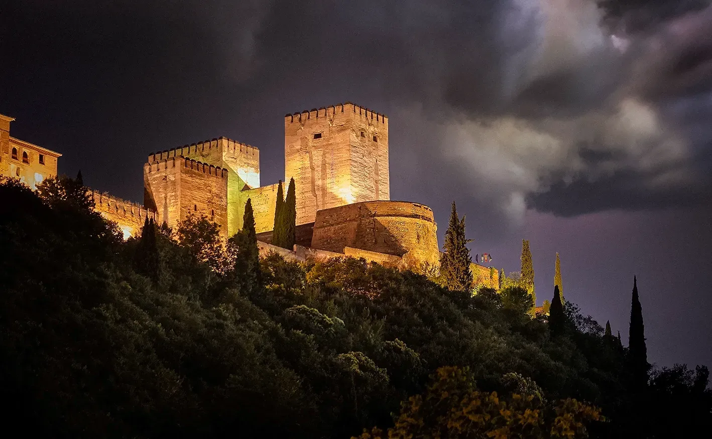 Lit-up Alhambra fortress at night, perched on a hill, against a dark, stormy sky.