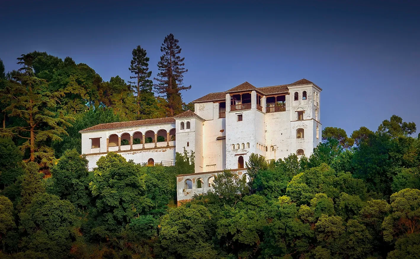 White building with arched windows atop a tree-filled hill under a blue sky.