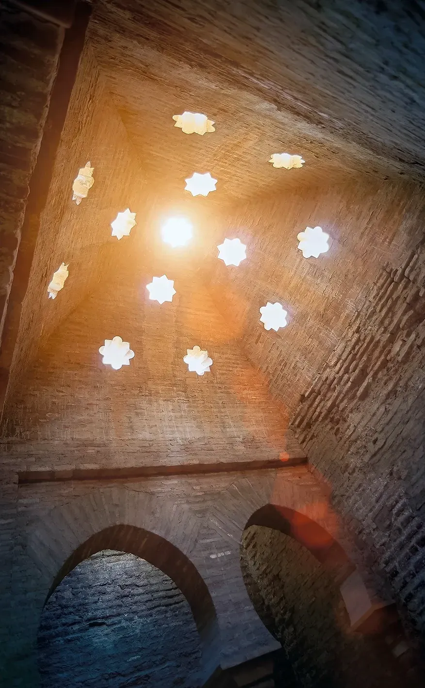 Brick ceiling with star-shaped skylights, sunlight streaming through. Arched doorway below.