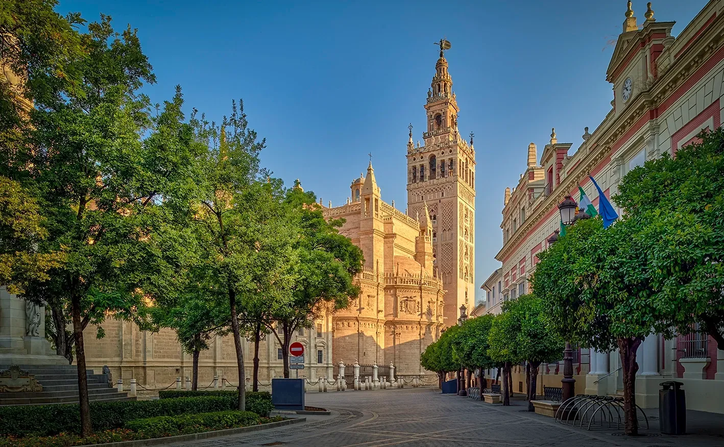 Seville Cathedral and Giralda tower on a sunny day. Orange trees line the square, buildings on the right.