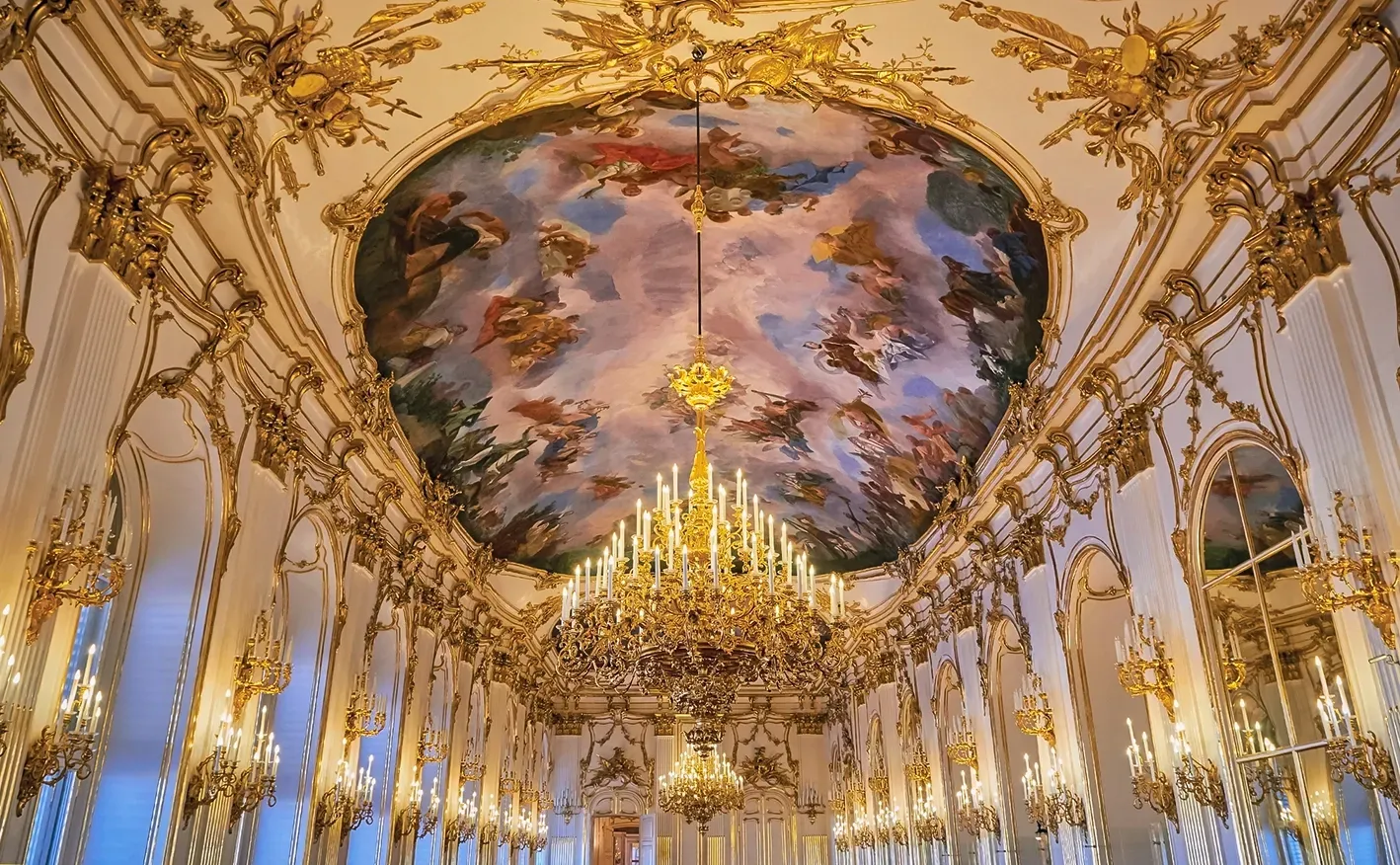Ornate ballroom ceiling with a painted mural, gold accents, and a large chandelier.