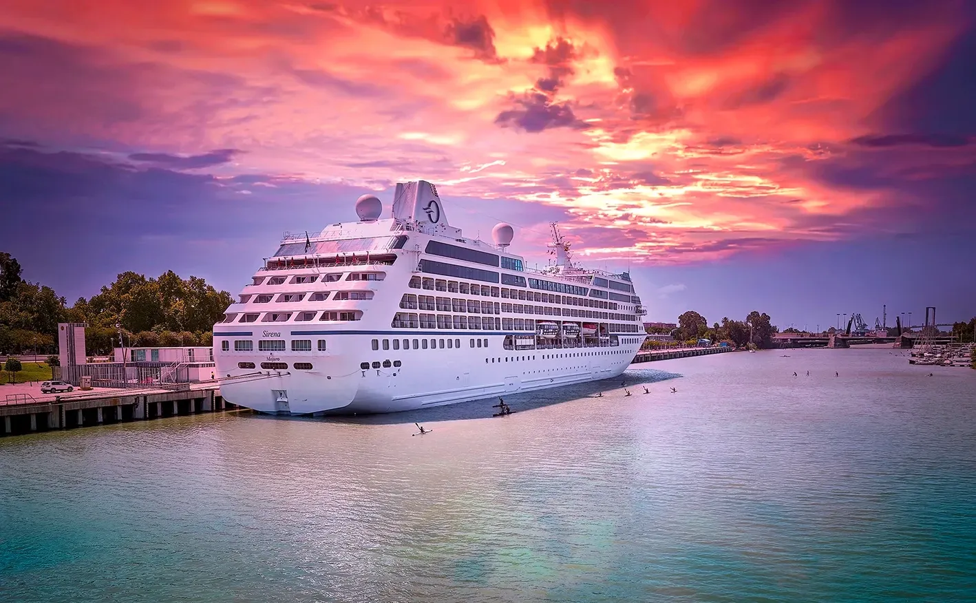 Cruise ship docked, water in foreground, vibrant sunset sky.