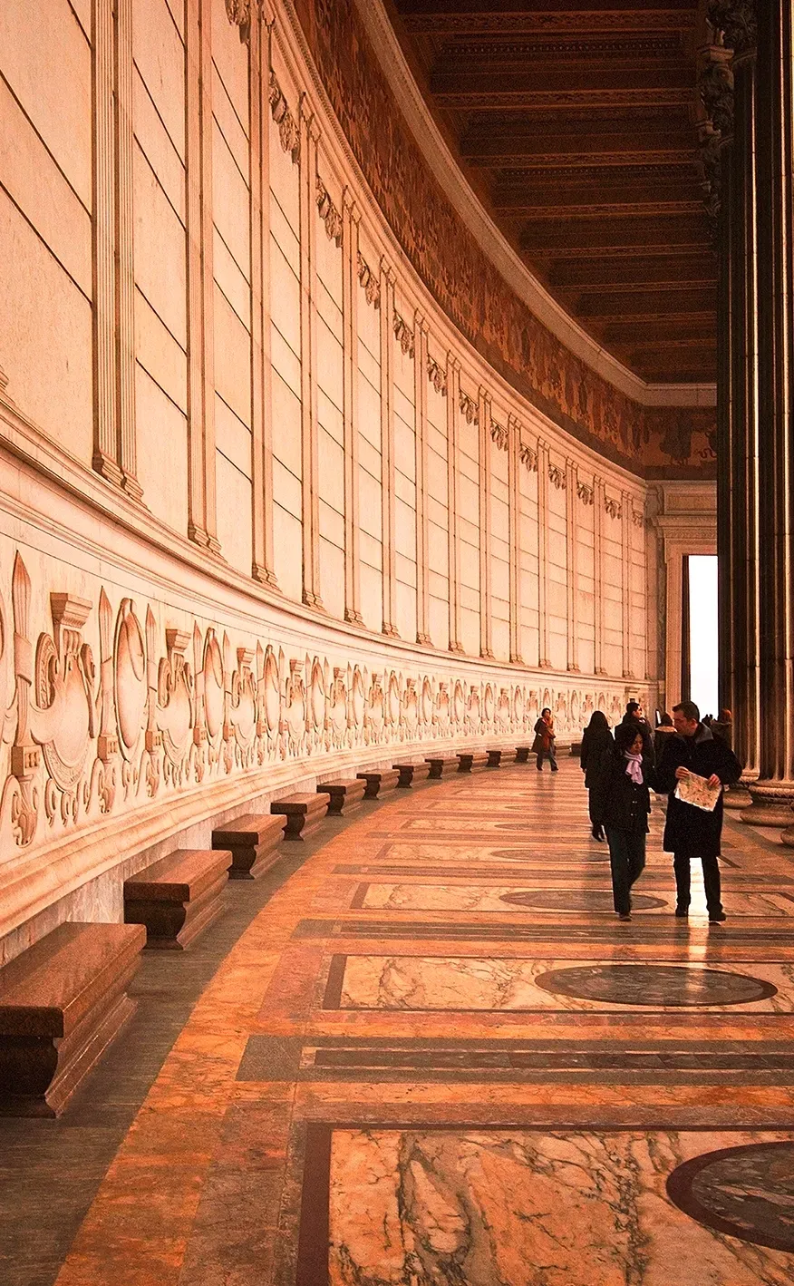 Interior hallway with carved wall, benches, patterned floor, and people walking.