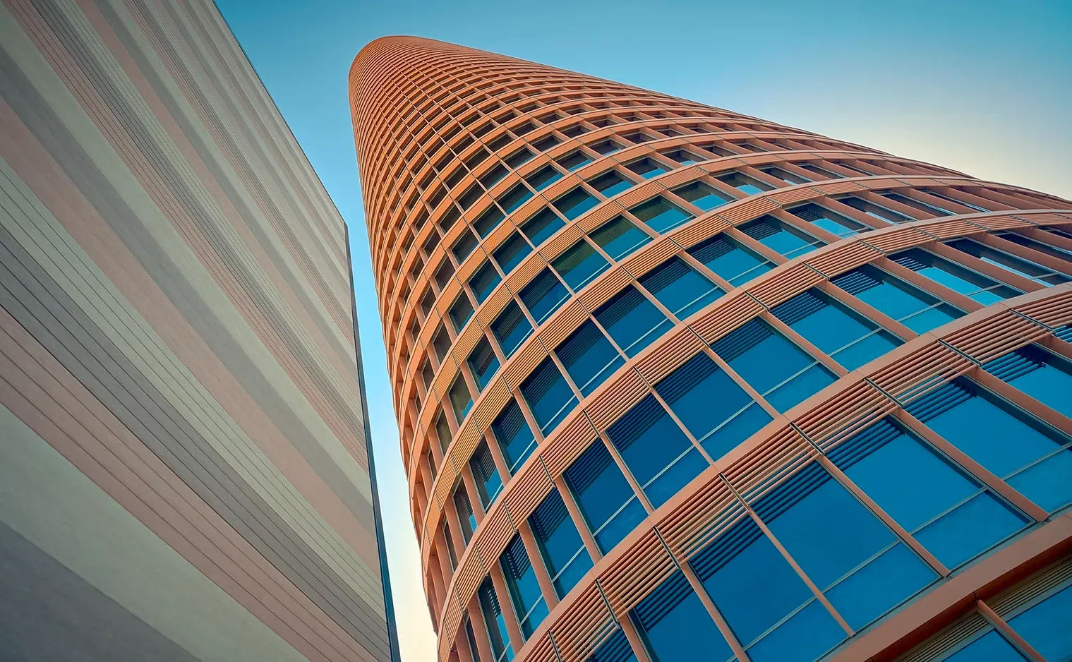 Skyscraper with rounded facade, blue windows, next to a striped building, blue sky.