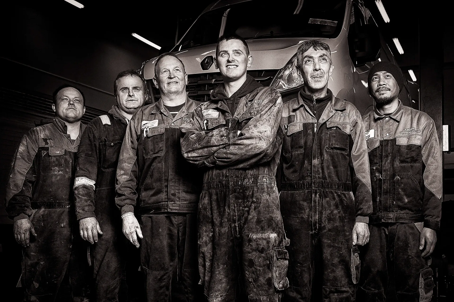 Six workers in dark, mud-covered coveralls pose together in a dimly lit garage space in front of a large vehicle.