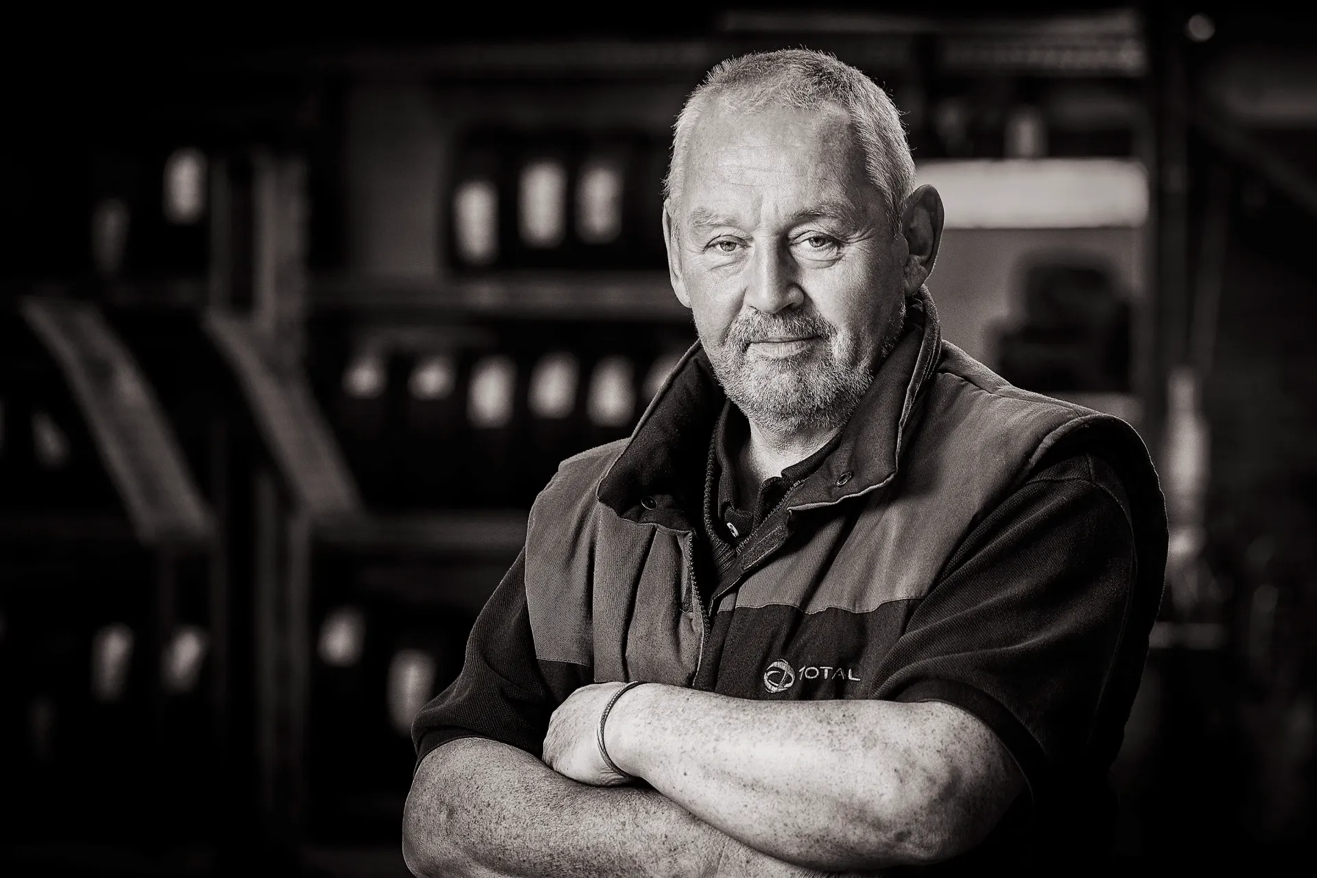 Man with arms crossed, wearing a vest, set in a workshop with tires in the background.