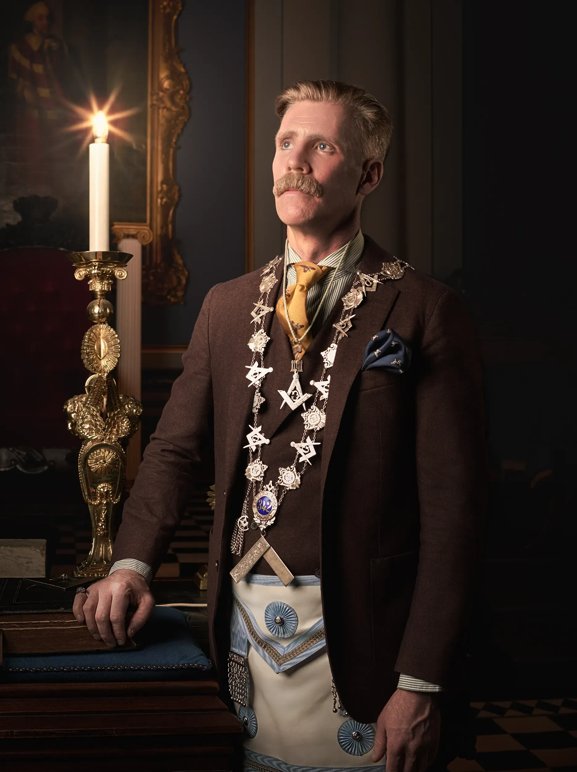 A person in a brown suit and Masonic apron, wearing a ceremonial collar, stands beside a lit candle in a dim, dark room.