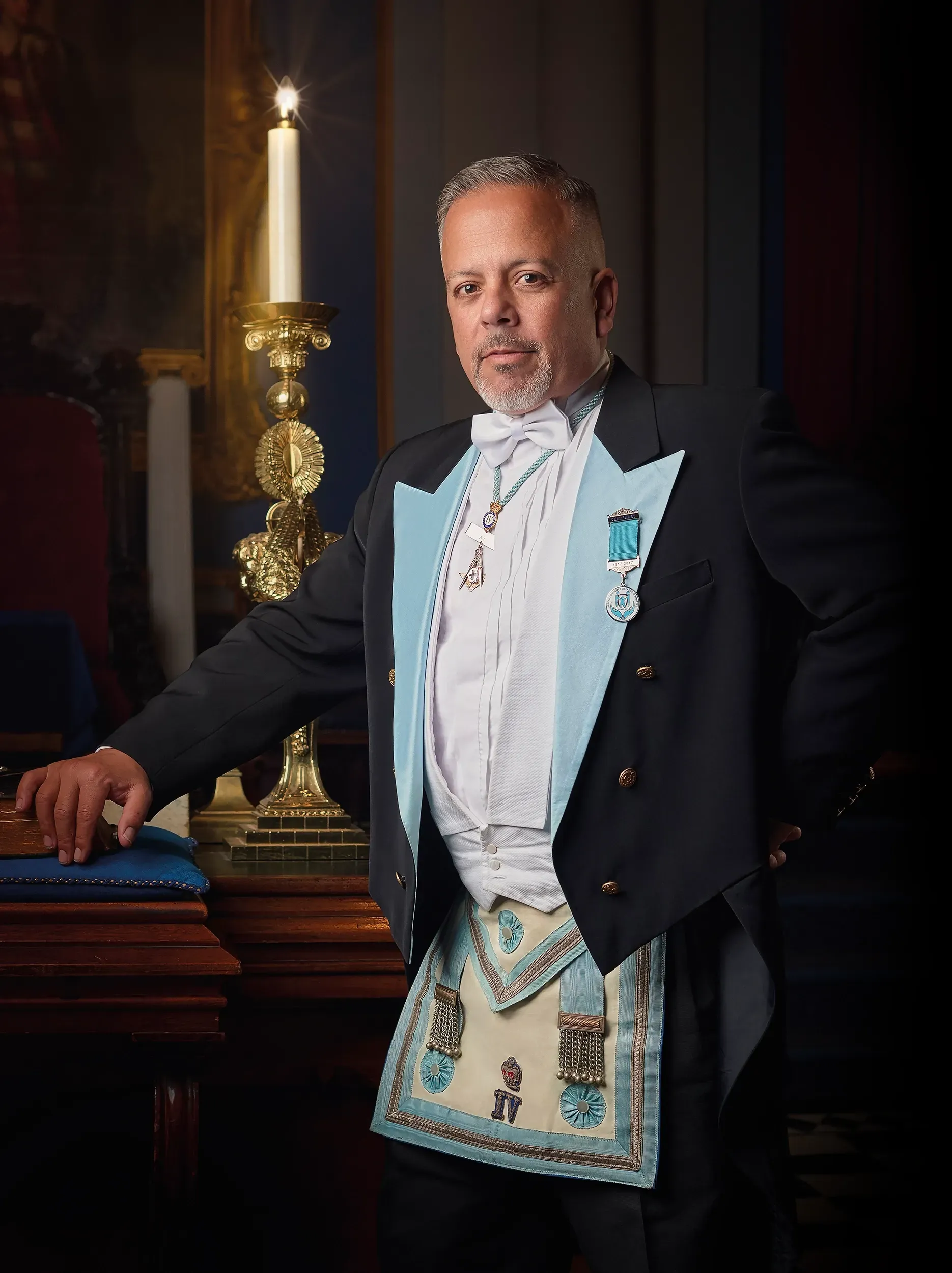 A person in formal Masonic attire with a light blue apron and collar standing next to a lit candle in a dark room.