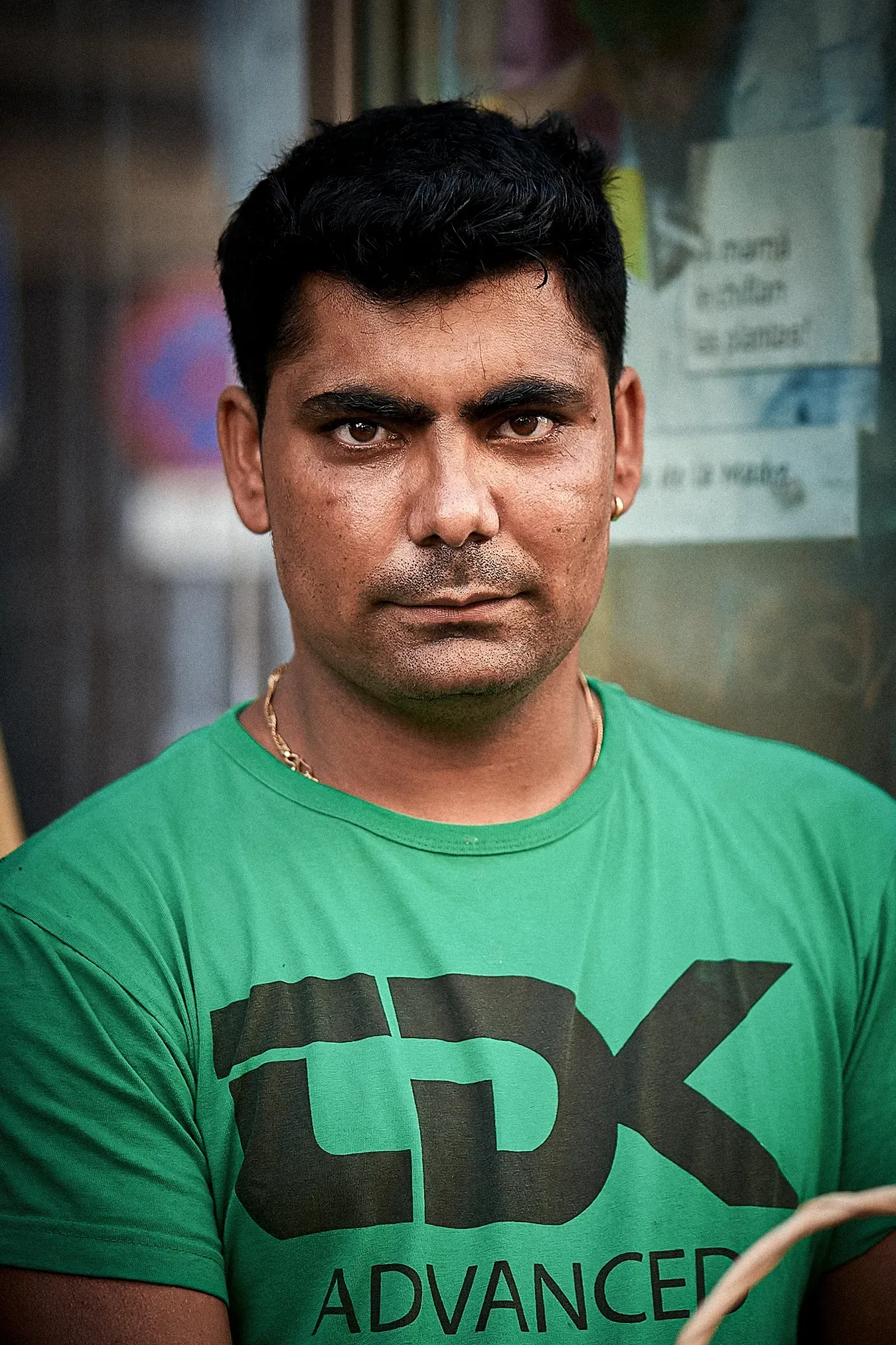 Man in green shirt with black text, looking directly at the camera. Outdoor setting, neutral expression.