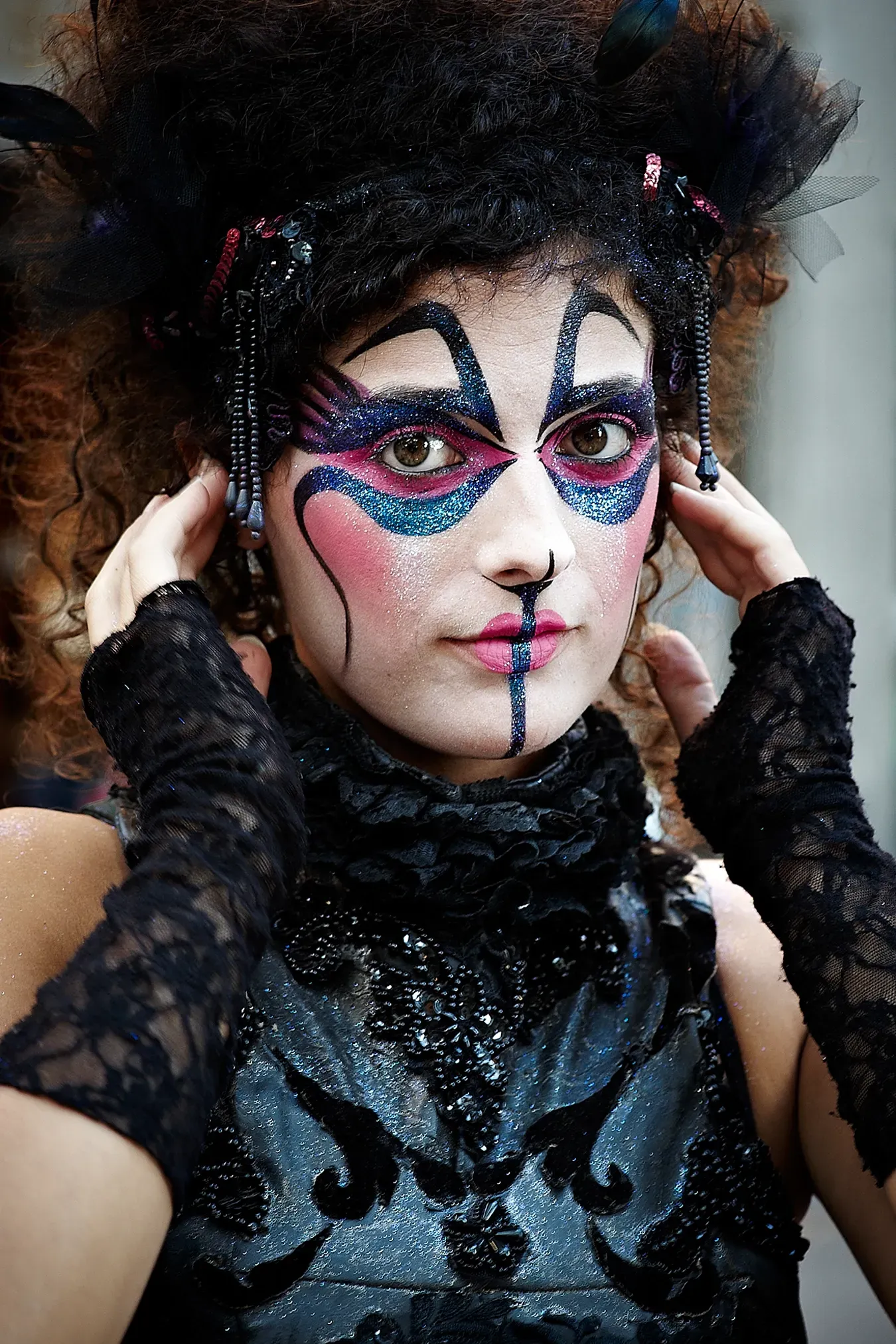 Woman with elaborate, colorful face paint and black costume, touching her temples.