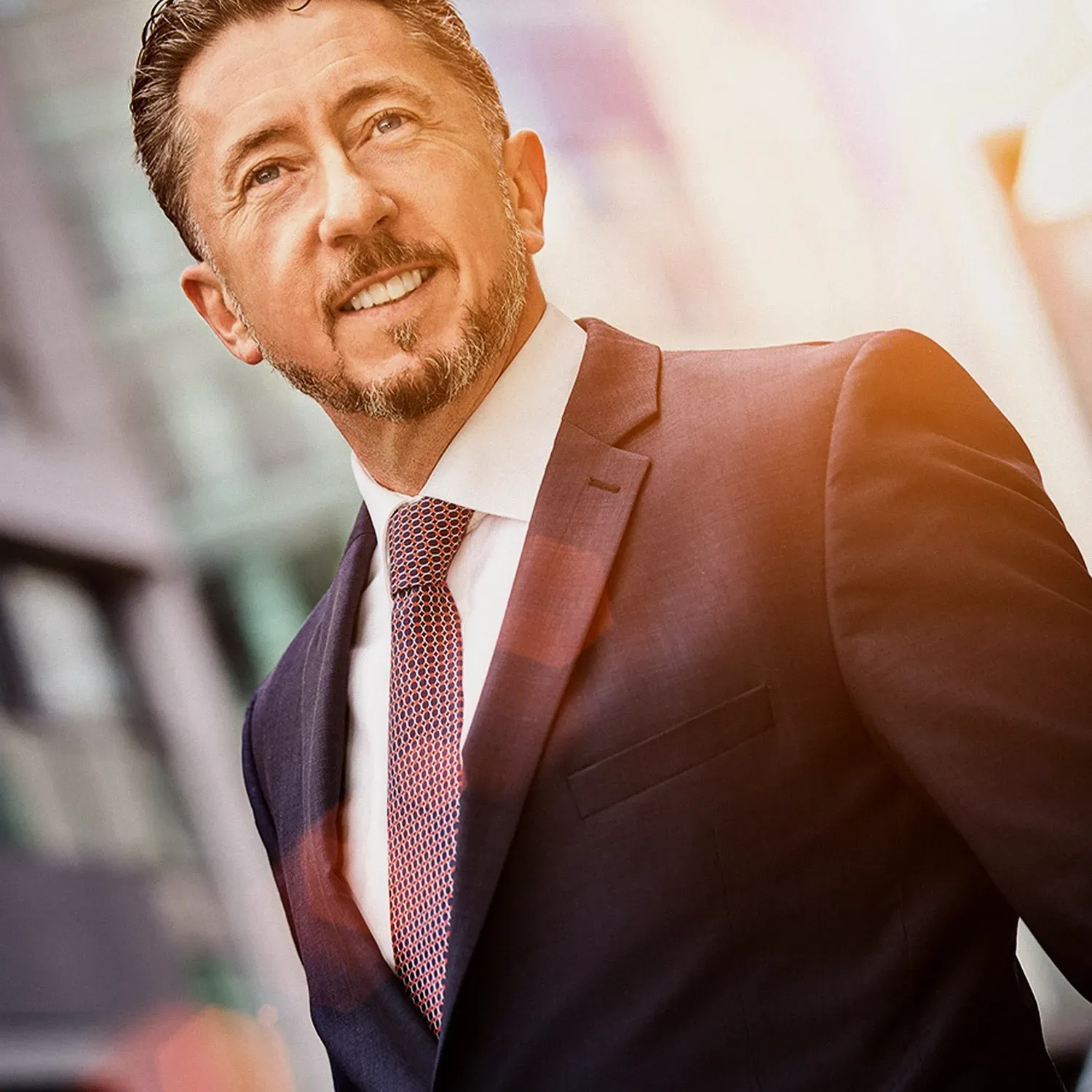 Man in a suit smiles, looking upward. Outdoors, blurred city background, warm lighting.