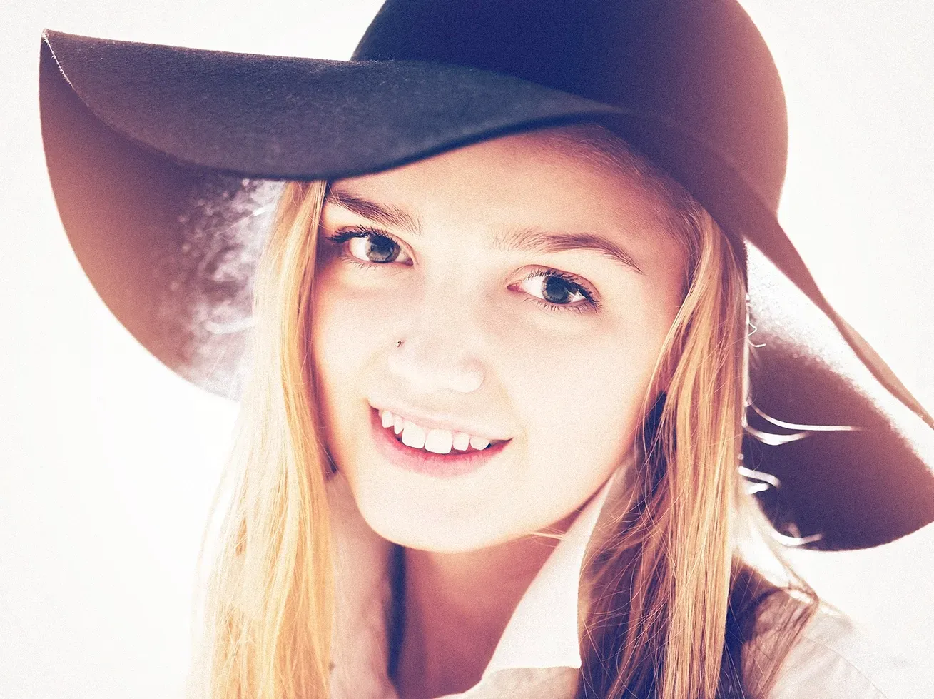 Woman with long blonde hair, smiling, wearing a wide-brimmed black hat and collared shirt.