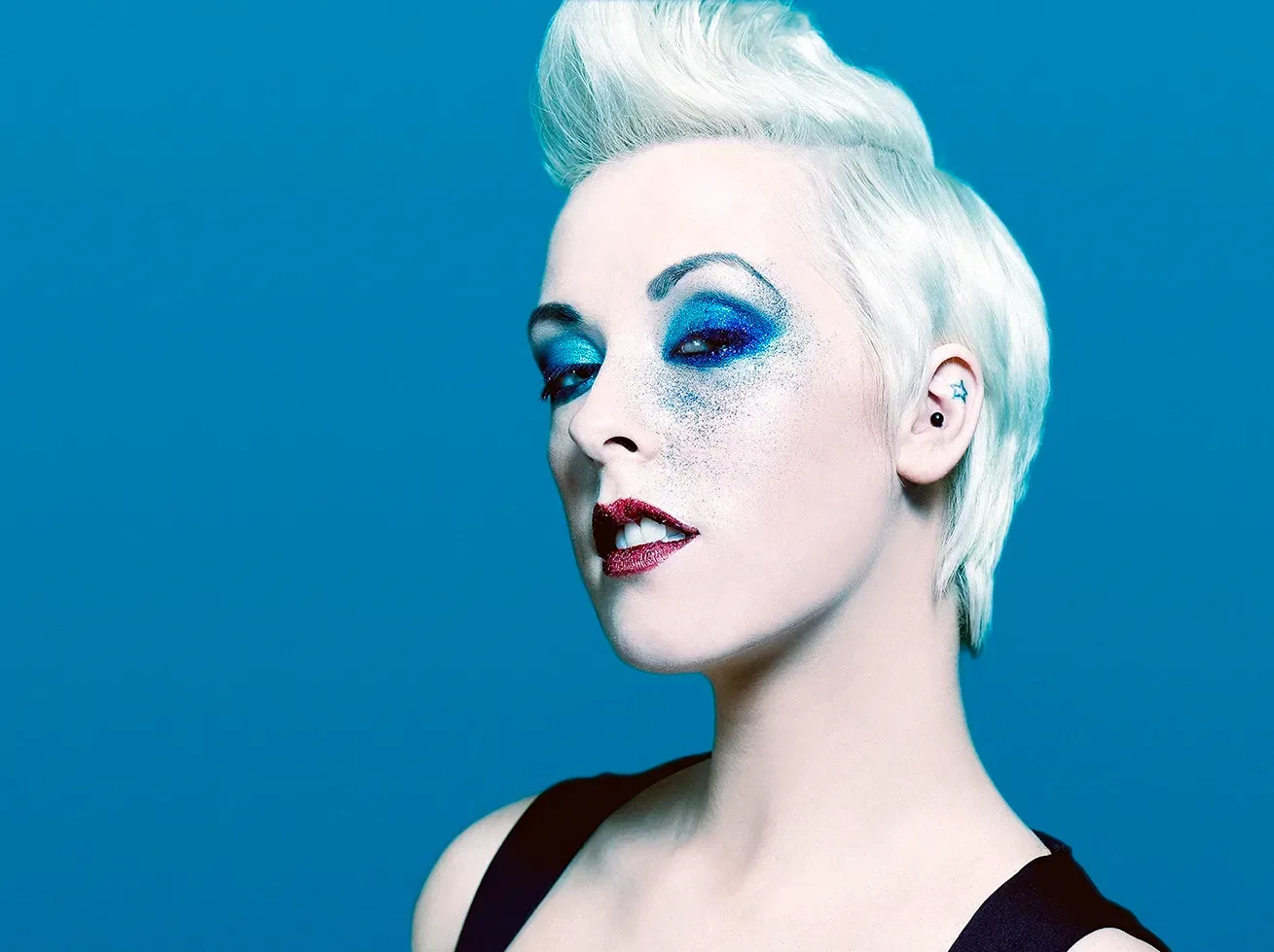 Woman with white hair styled upward, wearing blue eyeshadow and red lipstick. Blue background.
