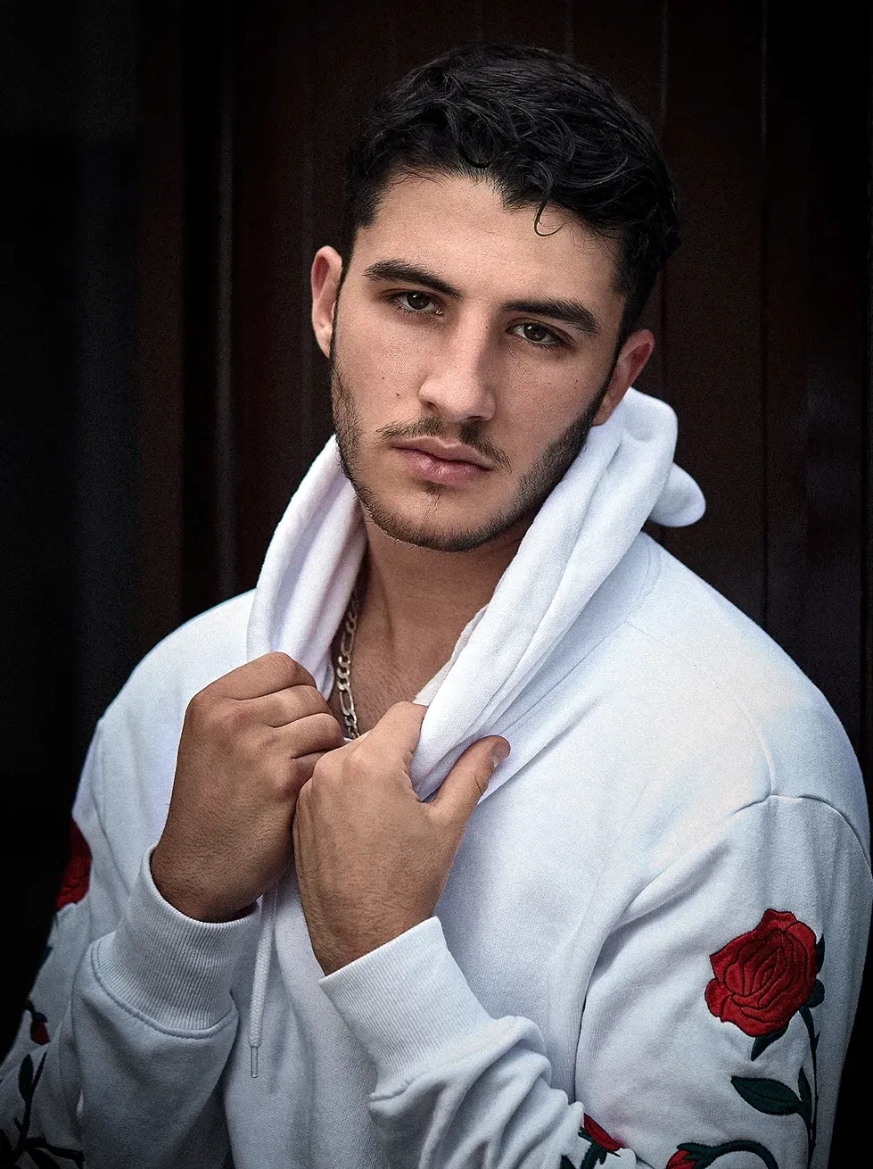 Man in white hoodie with rose accents, holding the hood, looking towards the viewer.