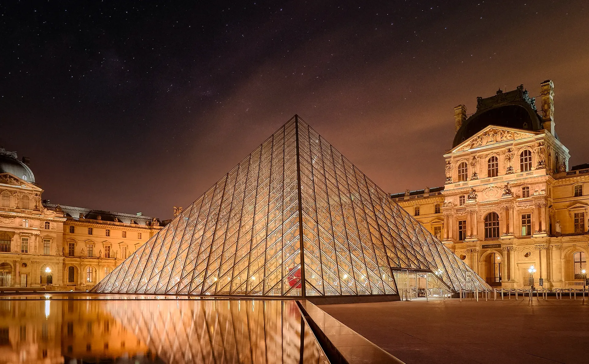 The Louvre Pyramid in Paris illuminated at night, reflected in a pool of water under a starry sky.