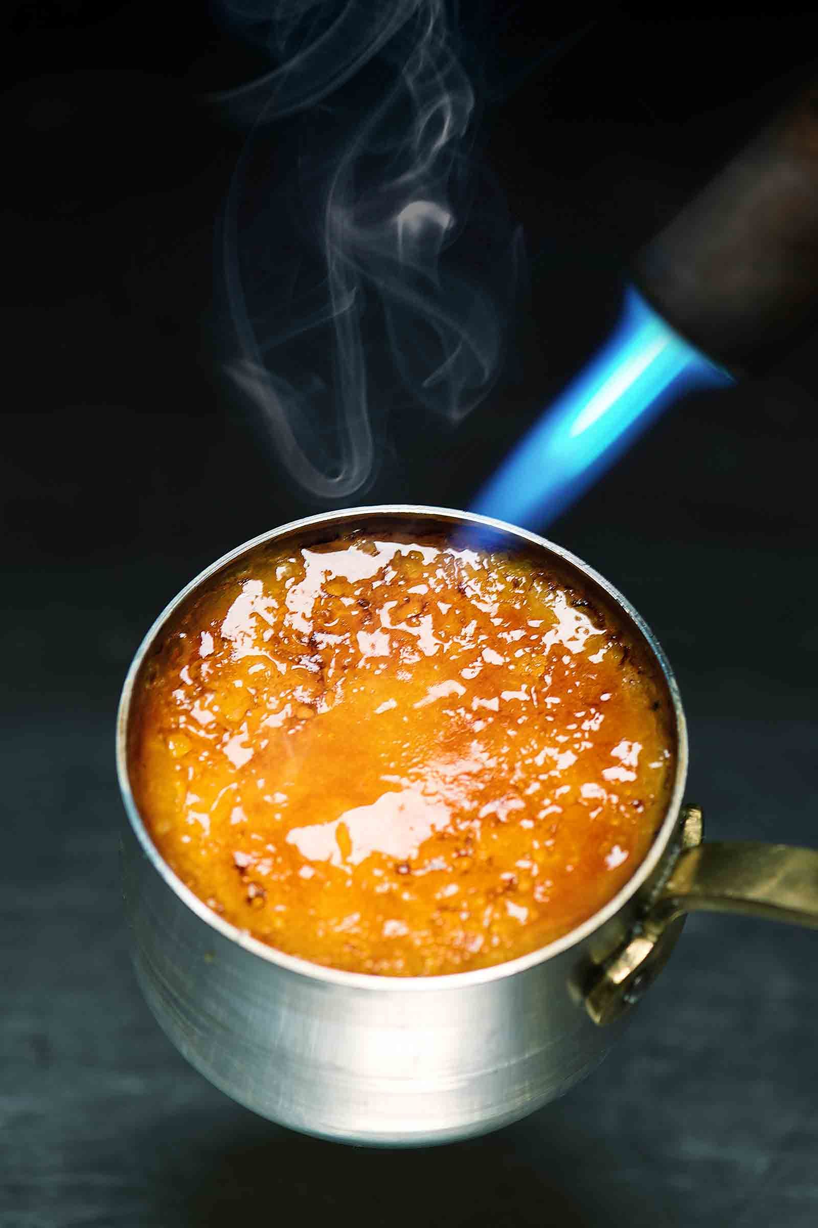 A small metal cup with bubbling, caramelized sugar being torched by a blue flame.