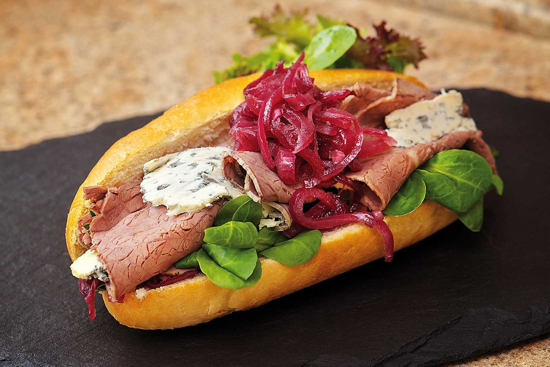 Roast beef sandwich with blue cheese, red onions, and greens on a roll, on a slate.