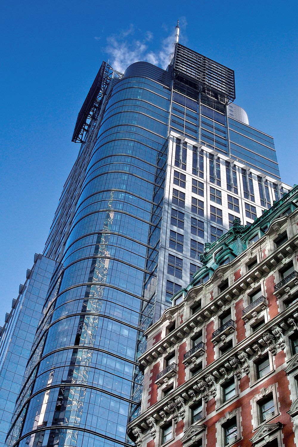 Modern glass skyscraper next to an ornate, older brick building, against a blue sky.
