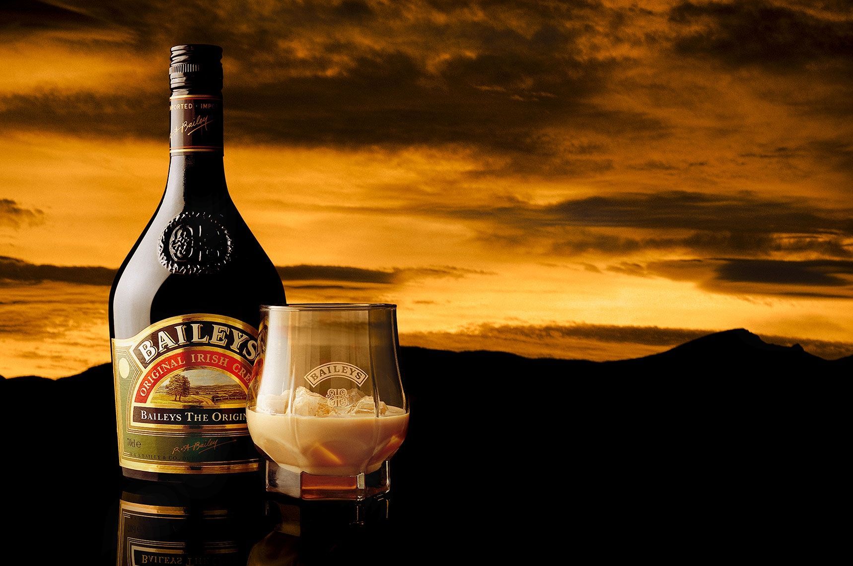 Bottle of Bailey's Irish Cream next to a filled glass. Sunset background with silhouetted mountains.