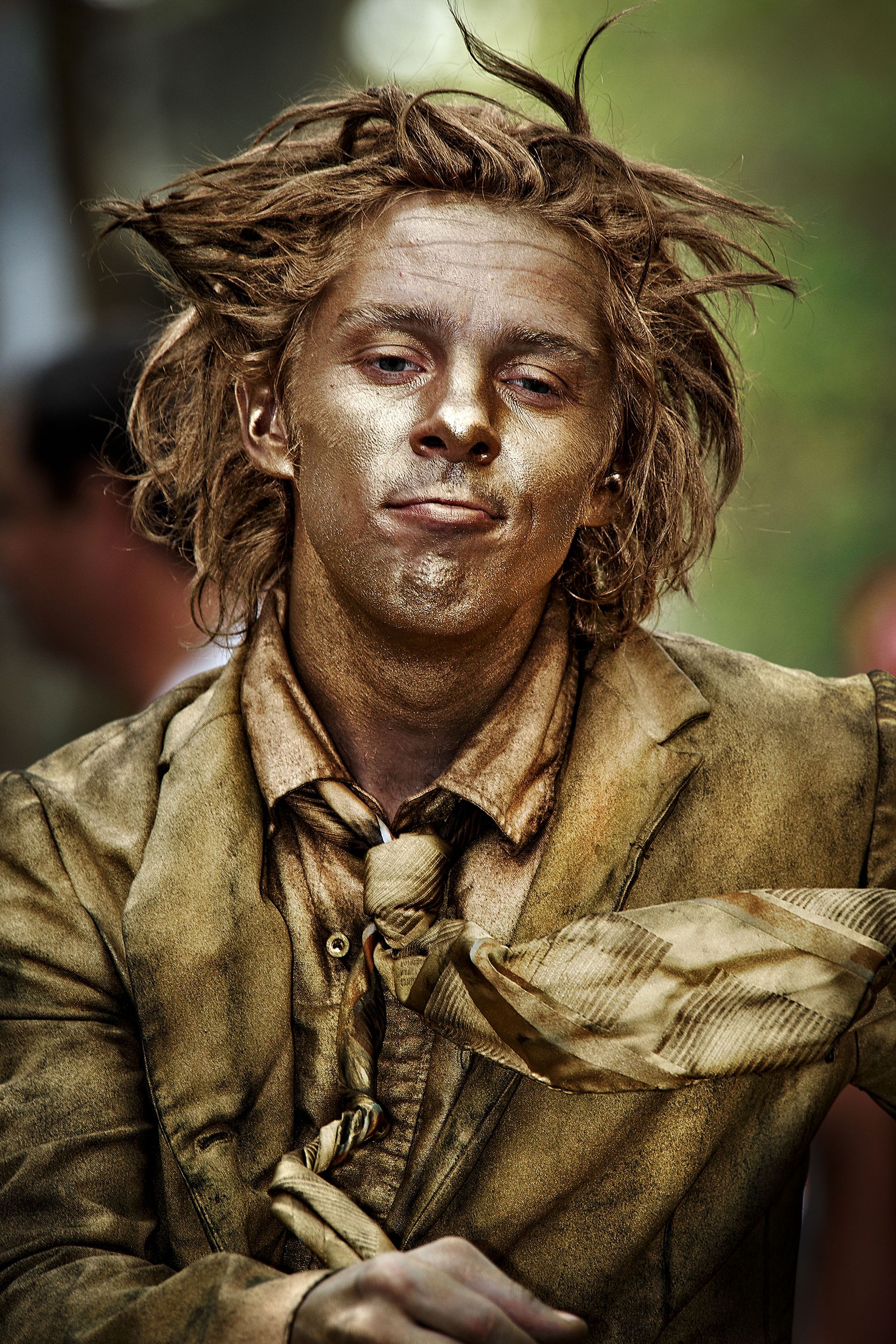 Man with gold body paint, messy hair, wearing suit, making a slightly smug face.