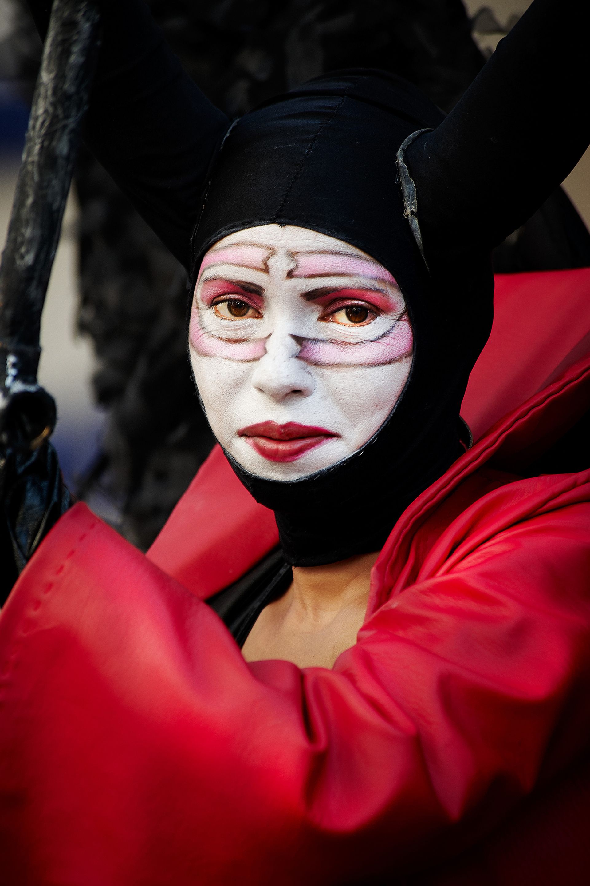 Person in black hood and red cloak with white face paint, red lips.