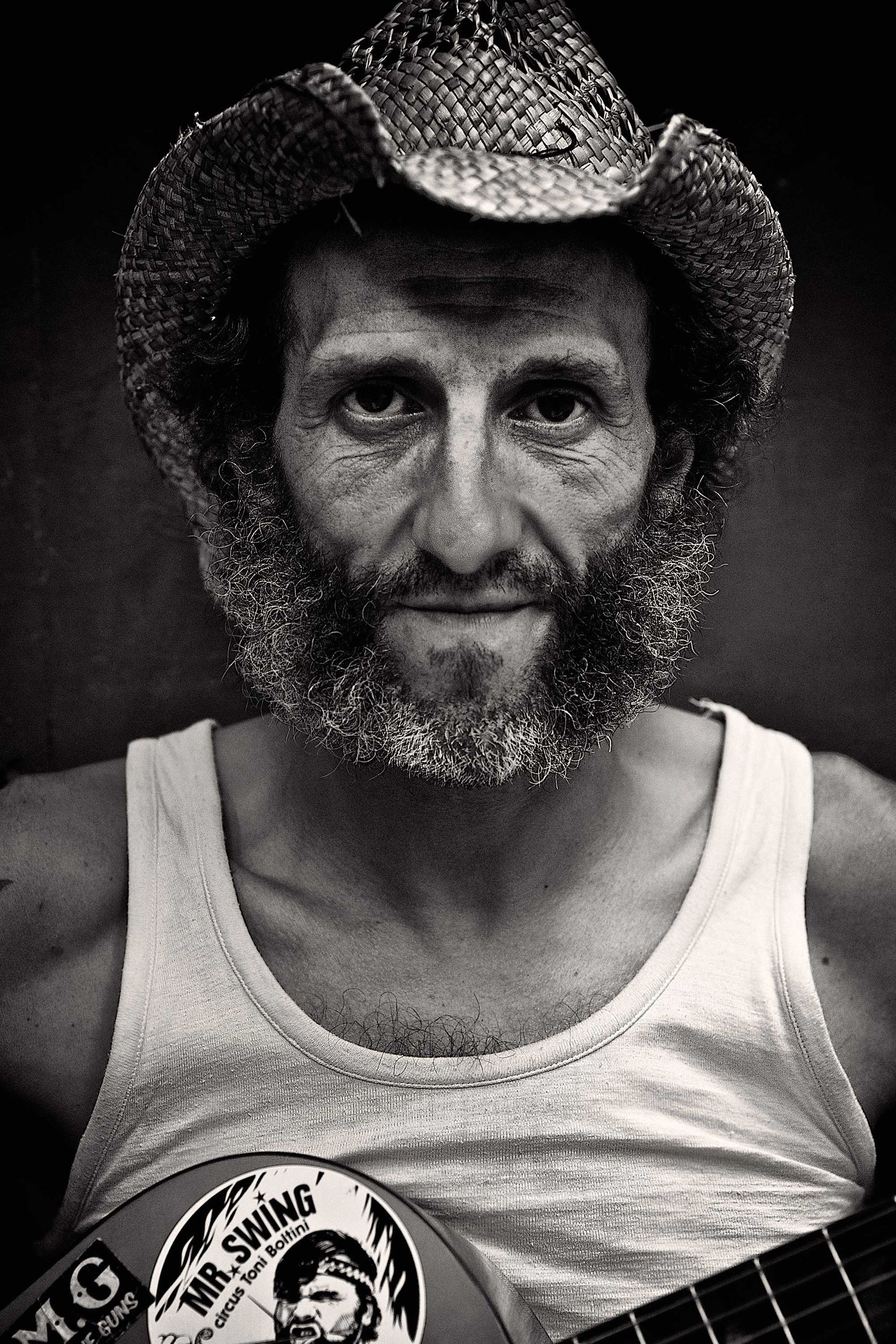 Man with beard and straw hat, wearing a tank top, holding a guitar with stickers, black and white.