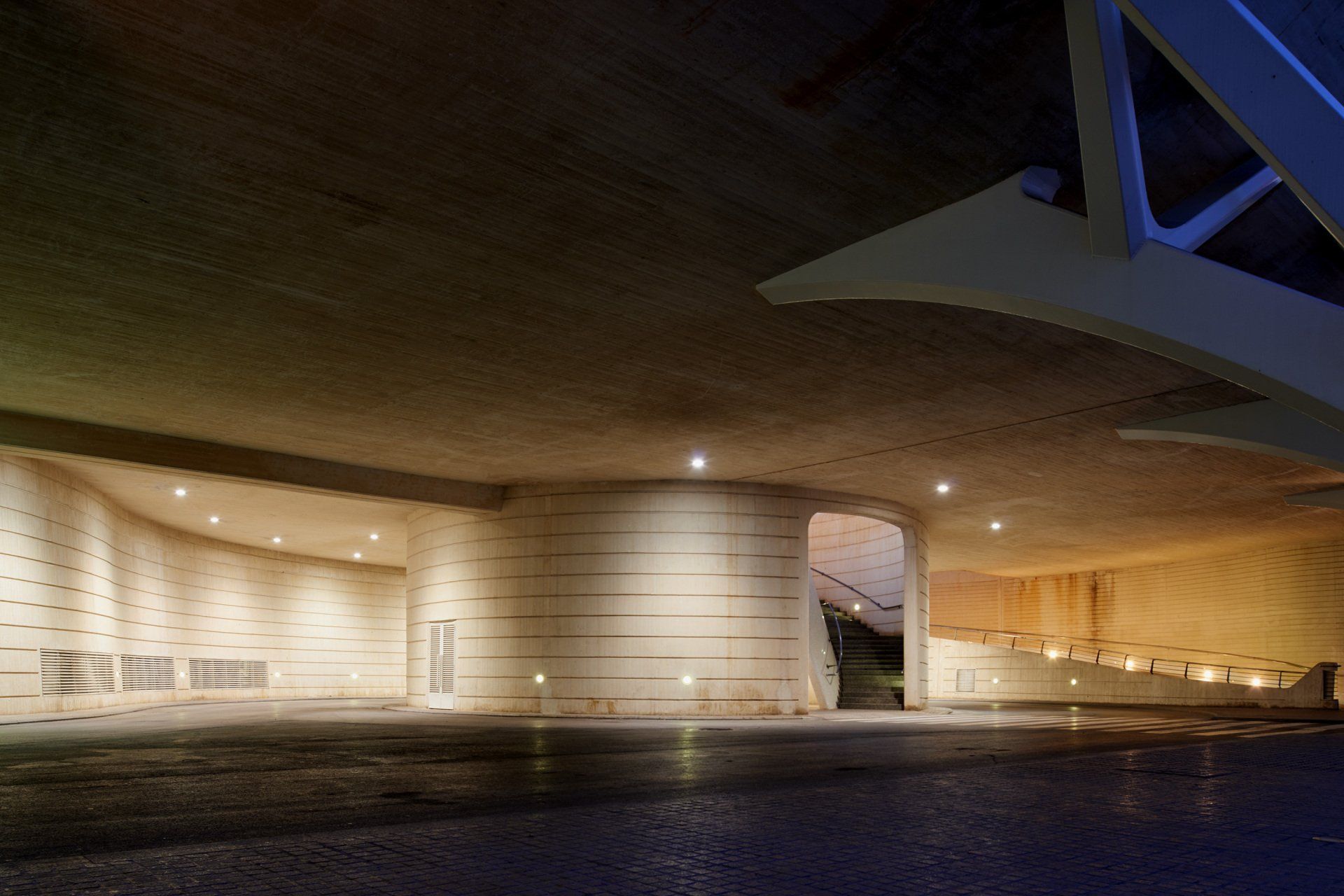 An illuminated, curved concrete underpass with layered walls, recessed lighting, and a paved walkway at night.