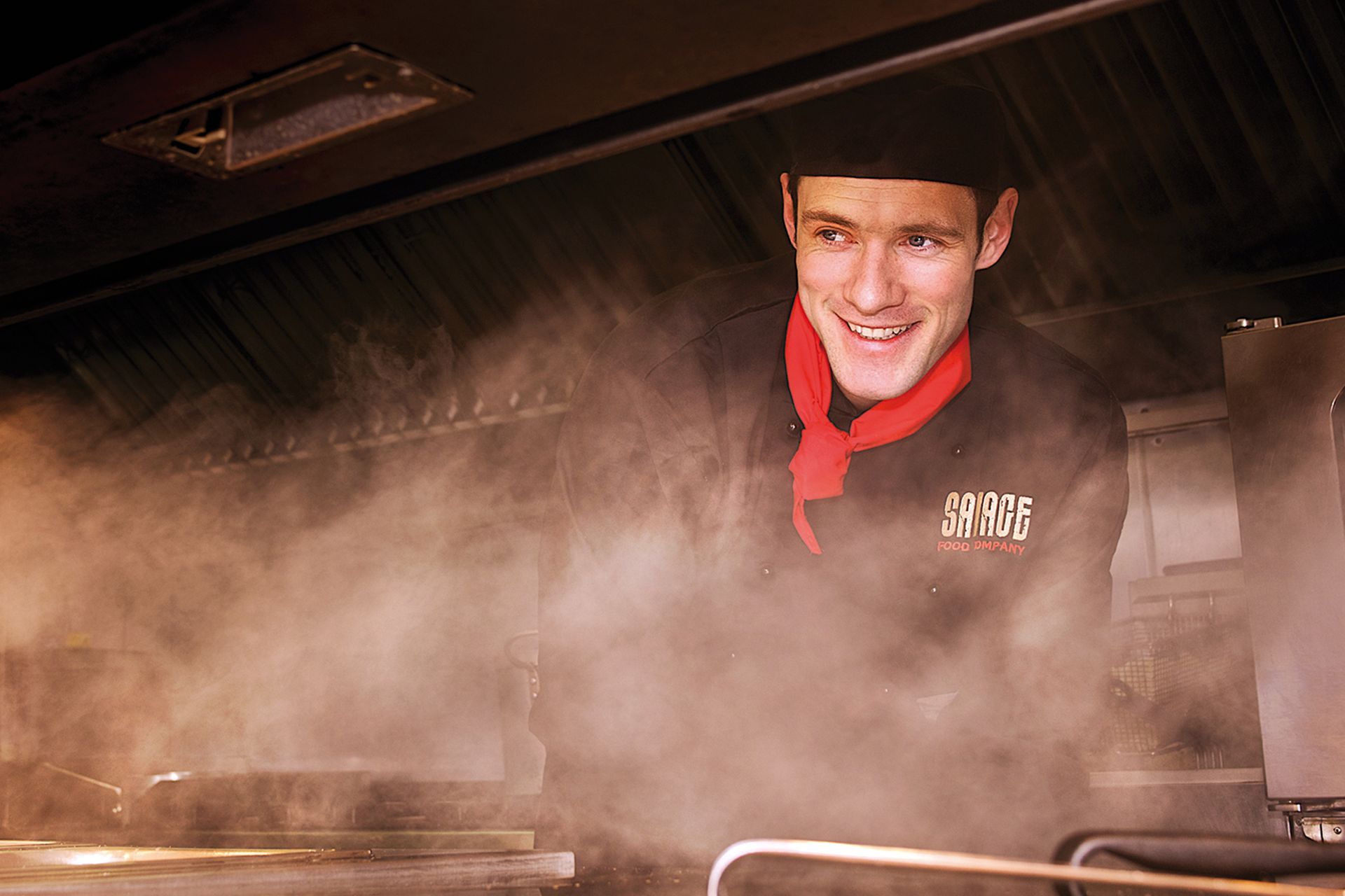 Chef smiling, amid steam in a restaurant kitchen, wearing black uniform and red neckerchief.