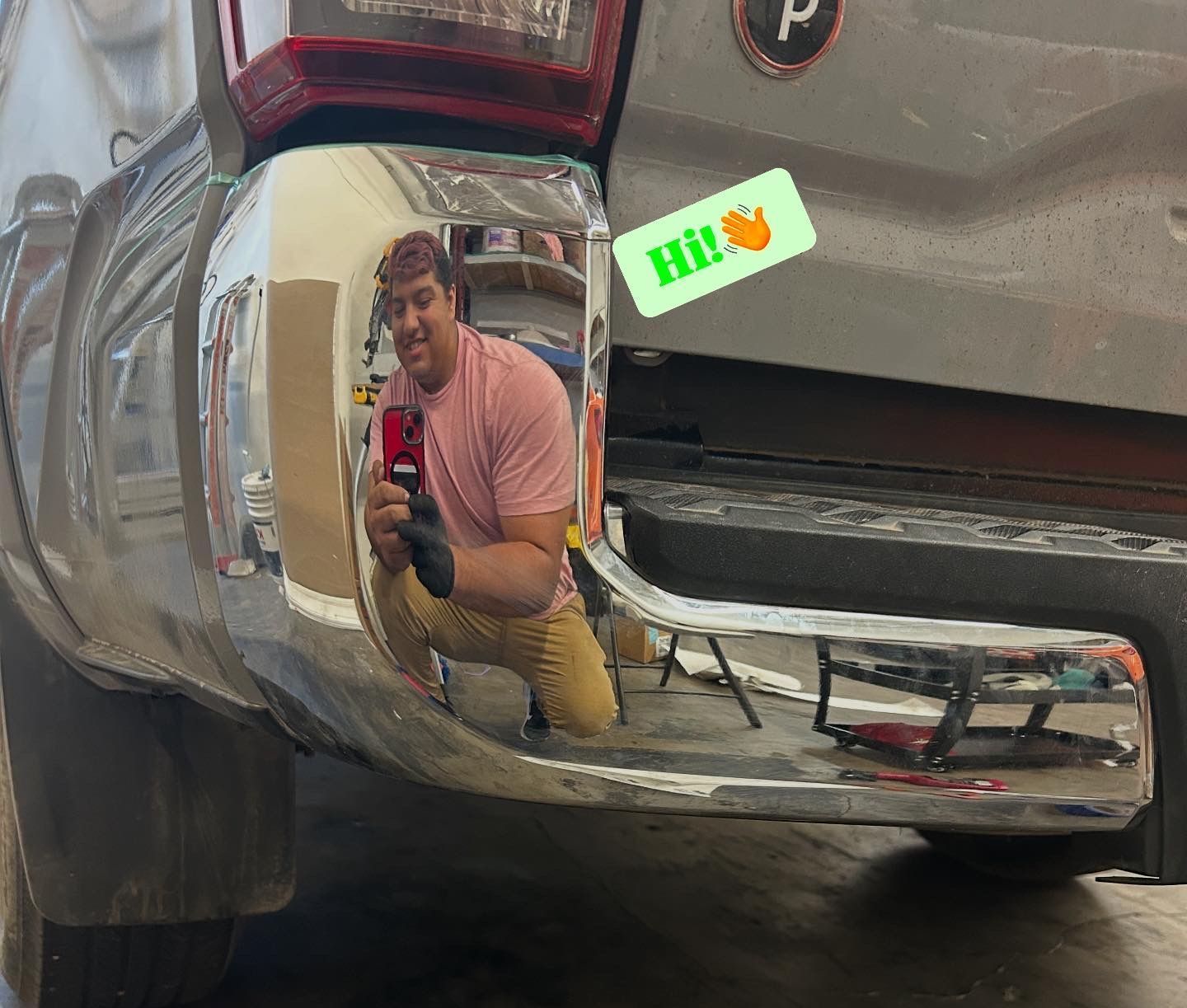 Person taking a mirror selfie in a shiny chrome truck bumper, with a small “Hi!” sticker nearby.