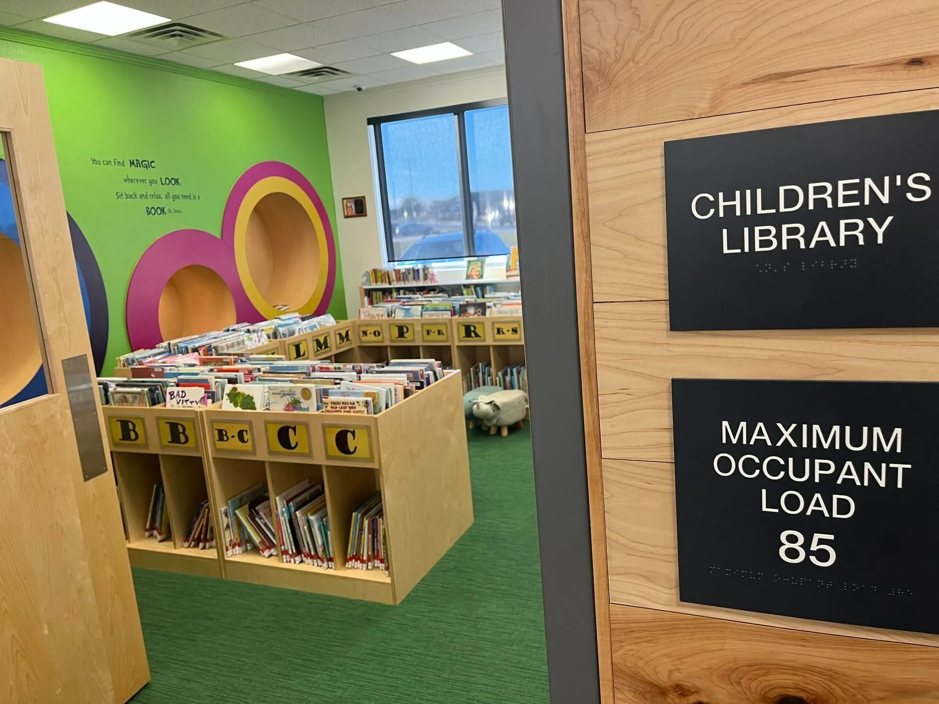 Children's library interior with bookshelves, colorful wall, and signage showing maximum occupant load of 85.