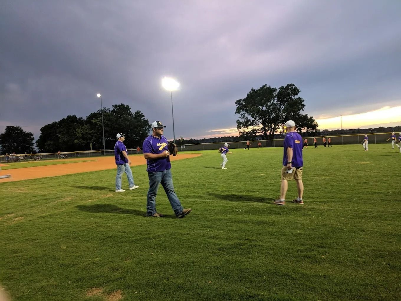 Men in purple shirts playing baseball on a green field under a cloudy sky.