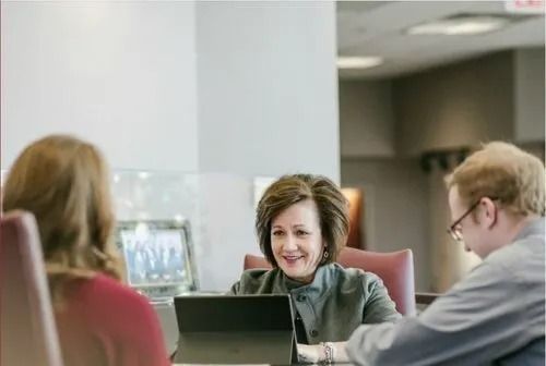 Three people in a meeting at a table. Woman in the center smiles, looking at a laptop.