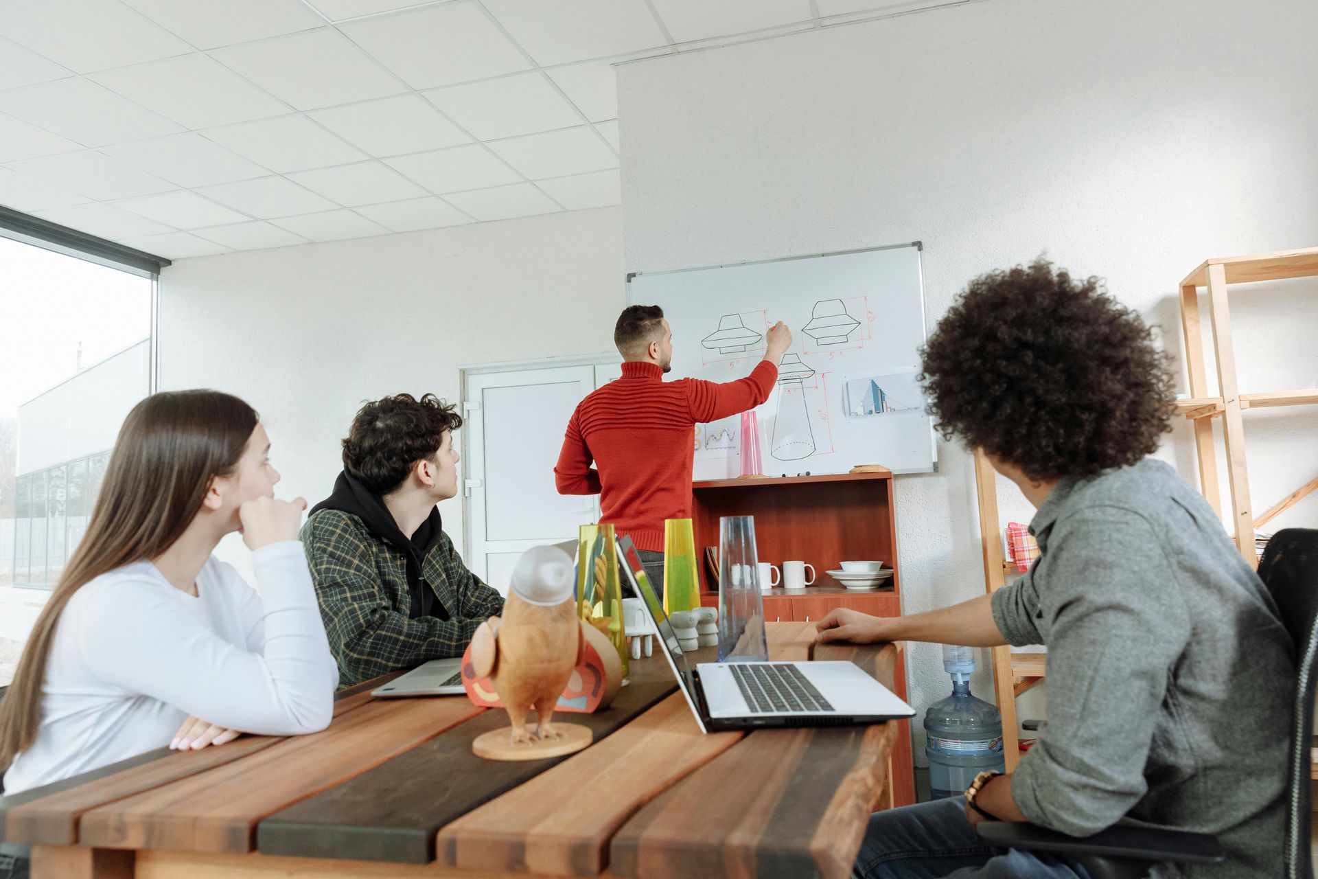 People in a meeting watching a presenter point at a whiteboard in a bright office
