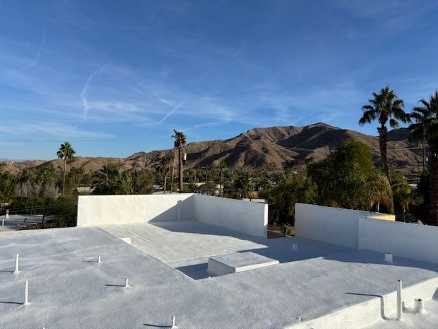 A white roof with low walls against a mountain backdrop. Palm trees dot the scene under a bright blue sky.