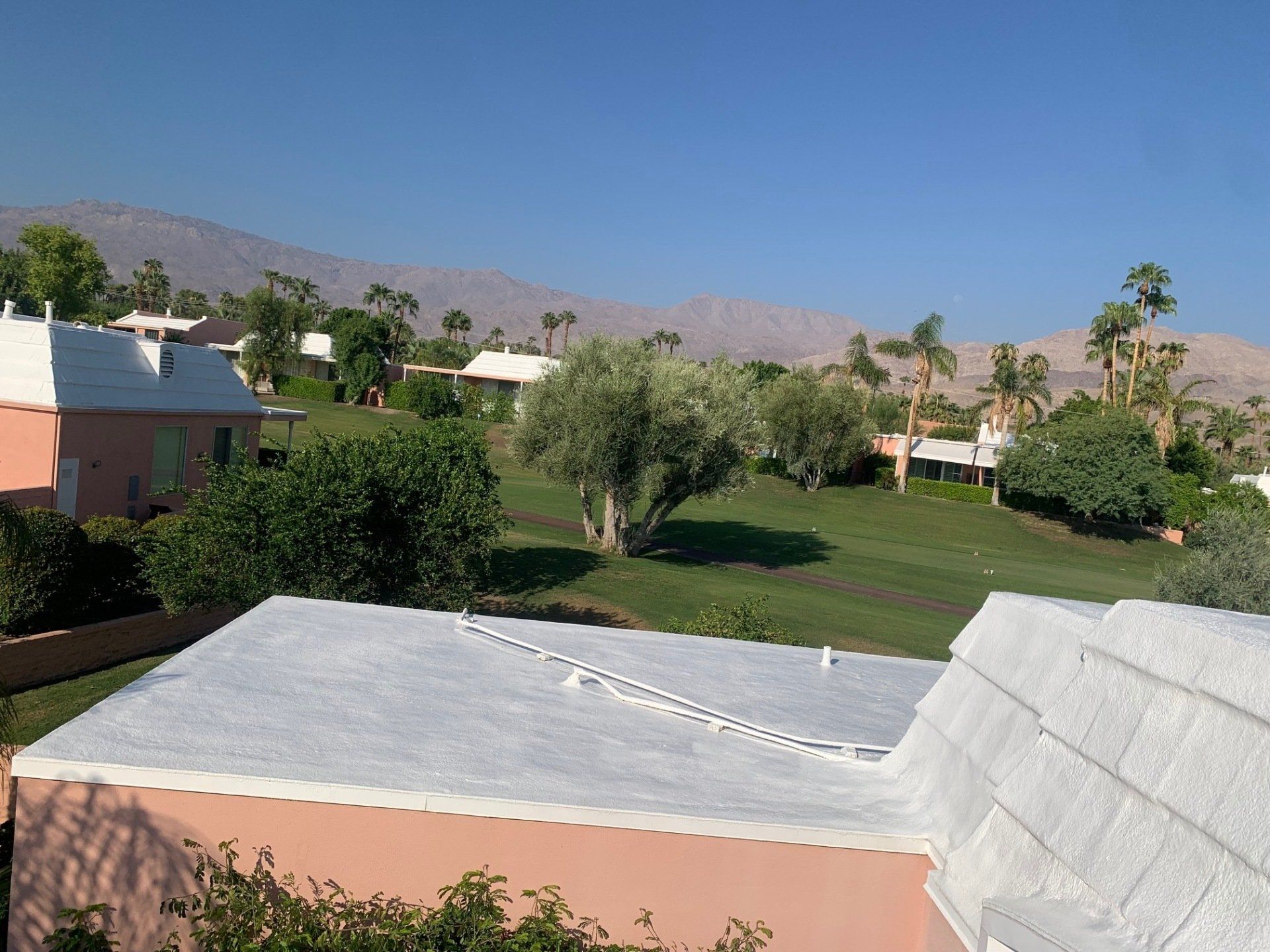 View of buildings with white and pink exteriors, a green lawn, trees, and mountains under a blue sky.