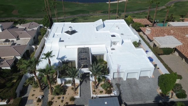 Aerial view of a large, white house with a flat roof, palm trees in the front, and a driveway leading to a three-car garage.