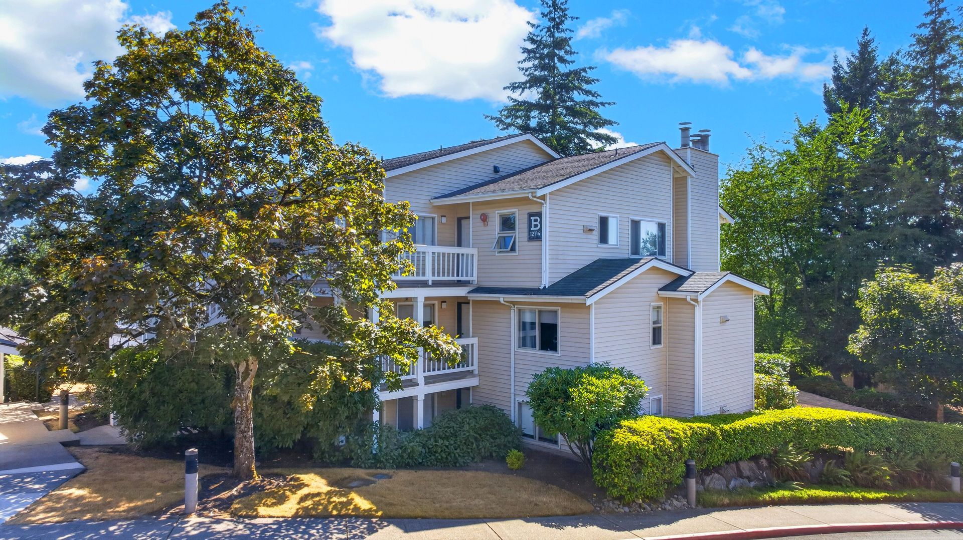 Two-story beige house with white trim, surrounded by trees and shrubs under a bright blue sky.