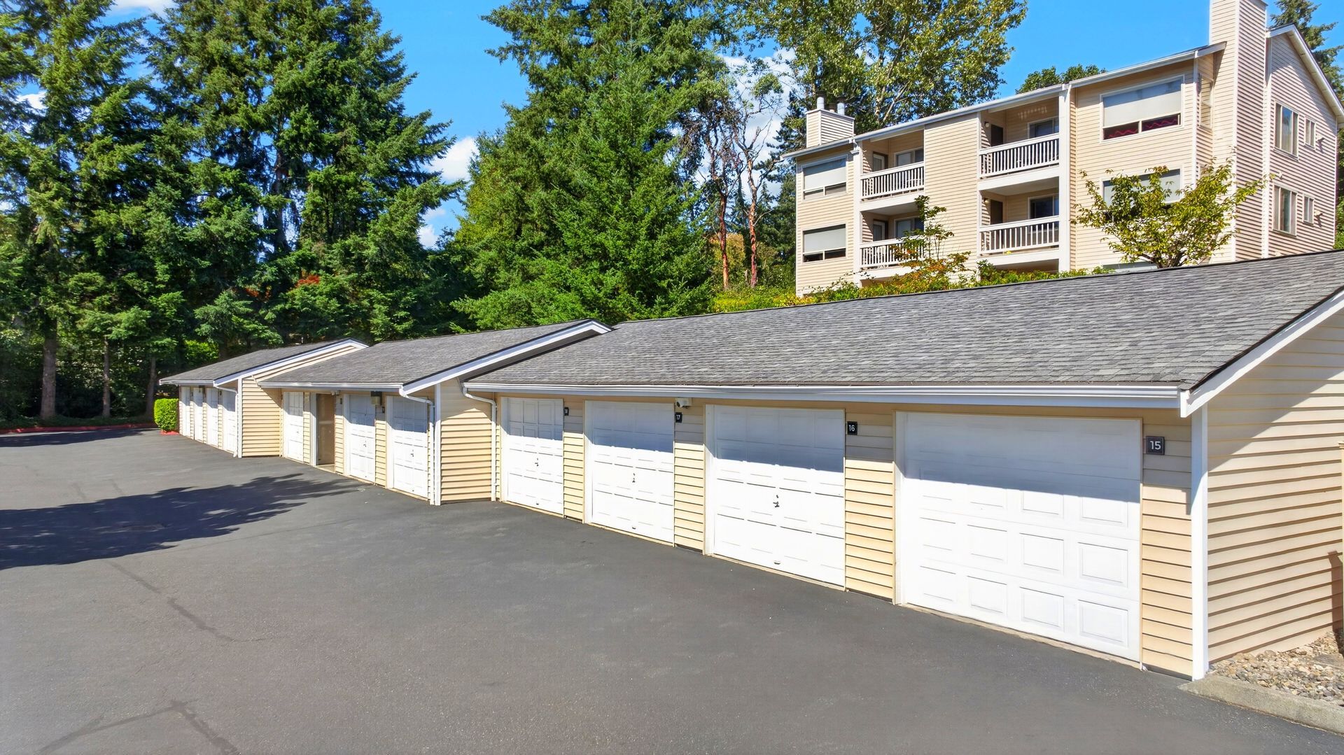 Row of beige garage units with white doors beside an asphalt road and apartment building in the background