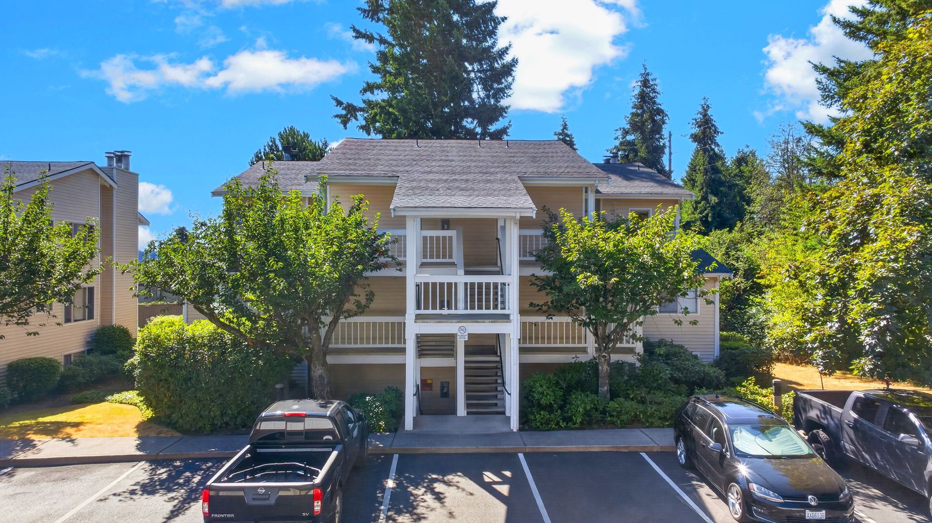 Apartment building with balconies, trees, and parked cars in a sunny lot
