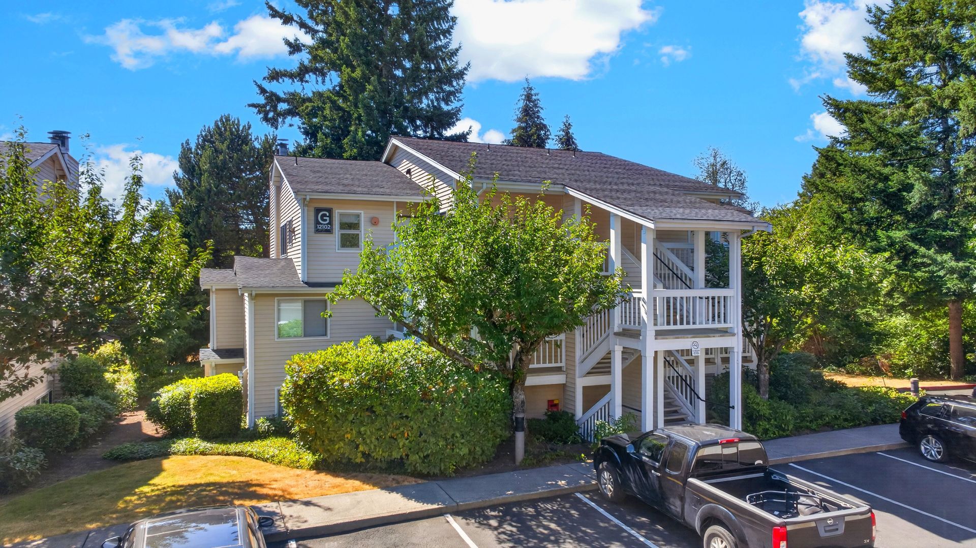 Two-story suburban house with white trim, green shrubs, and parked cars on a sunny day