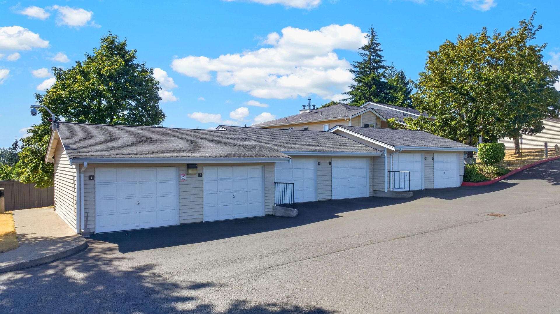 Row of white garage units beside a sloped street under a bright blue sky, with trees nearby