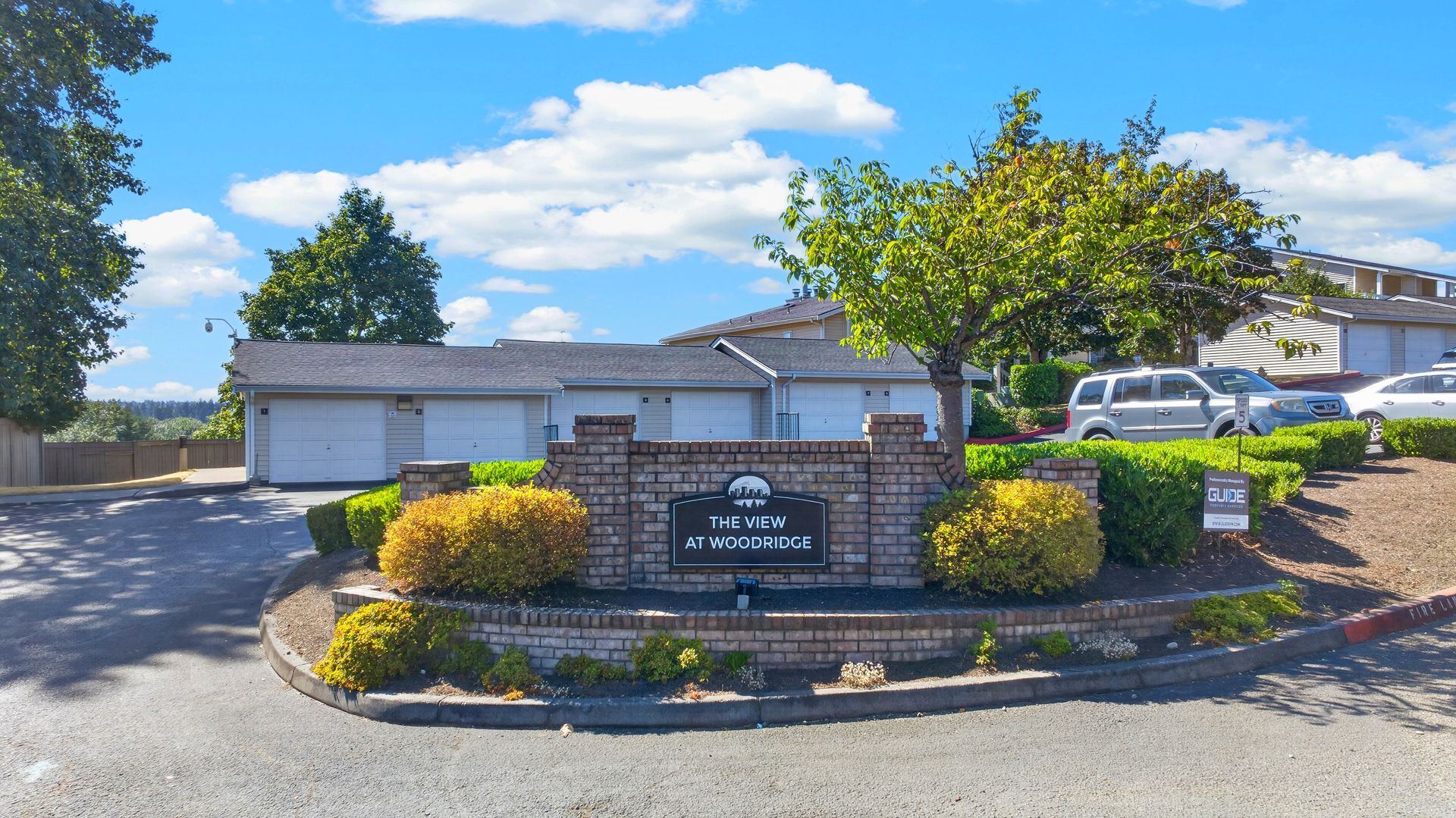 Landscaped entrance with stone sign, shrubs, and parked cars under a blue sky
