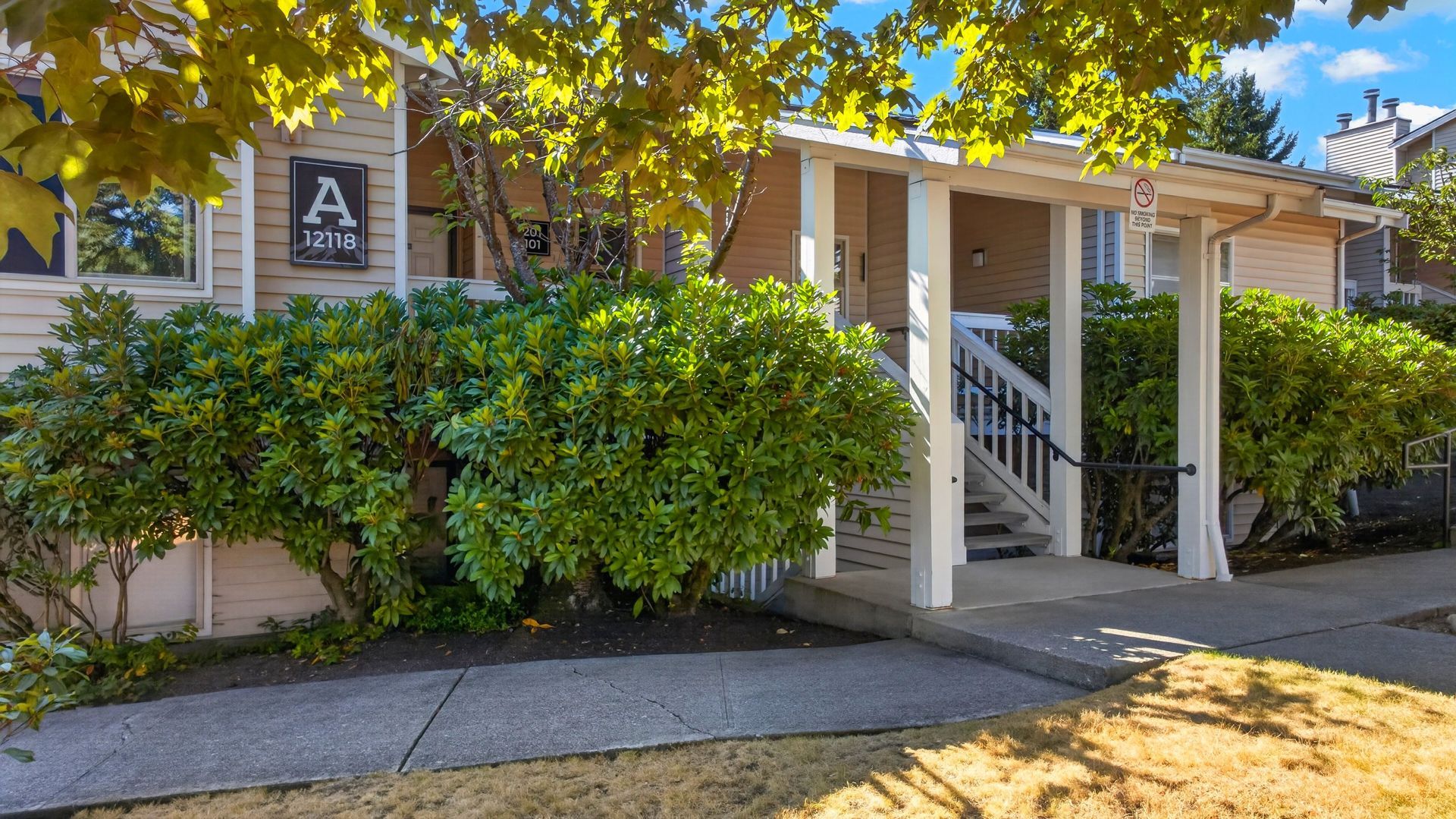 Apartment building entrance with white columns, stairs, and green shrubs under a sunny tree
